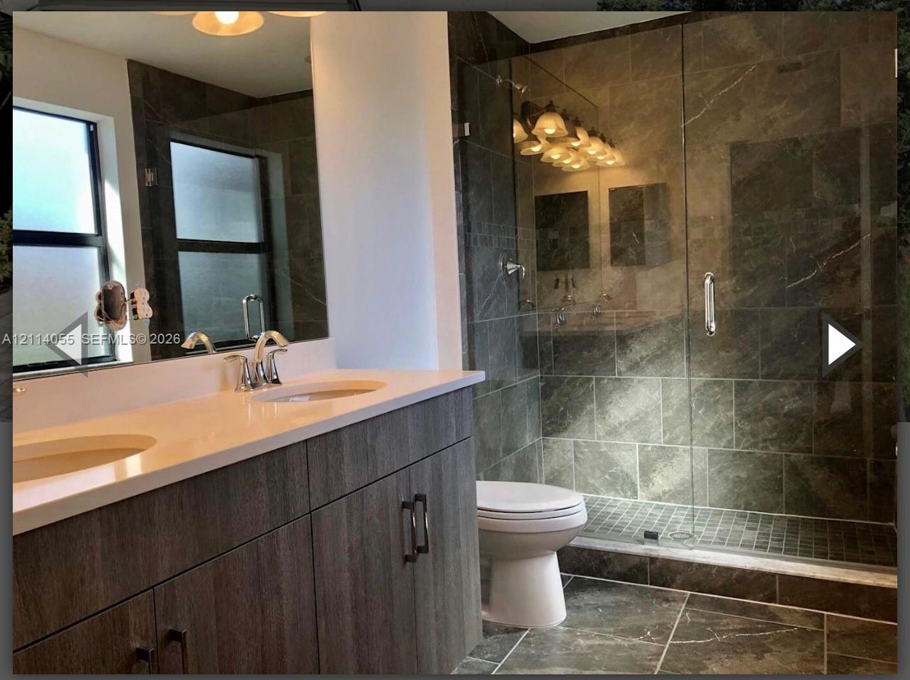 Dual-sink vanity with a white countertop and wood-finish cabinetry, walk-in shower with a glass enclosure, floor-to-ceiling tile work, and large-format floor tiling Dual-sink vanity with a white countertop and wood-finish cabinetry, walk-in shower with a glass enclosure, floor-to-ceiling tile work, and large-format floor tiling