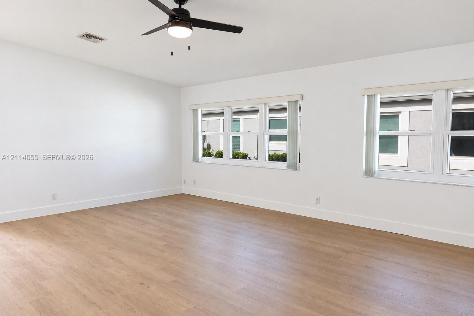 Neutral-toned interior space featuring wood-finish flooring, dual windows, and a matte black ceiling fan with integrated lighting