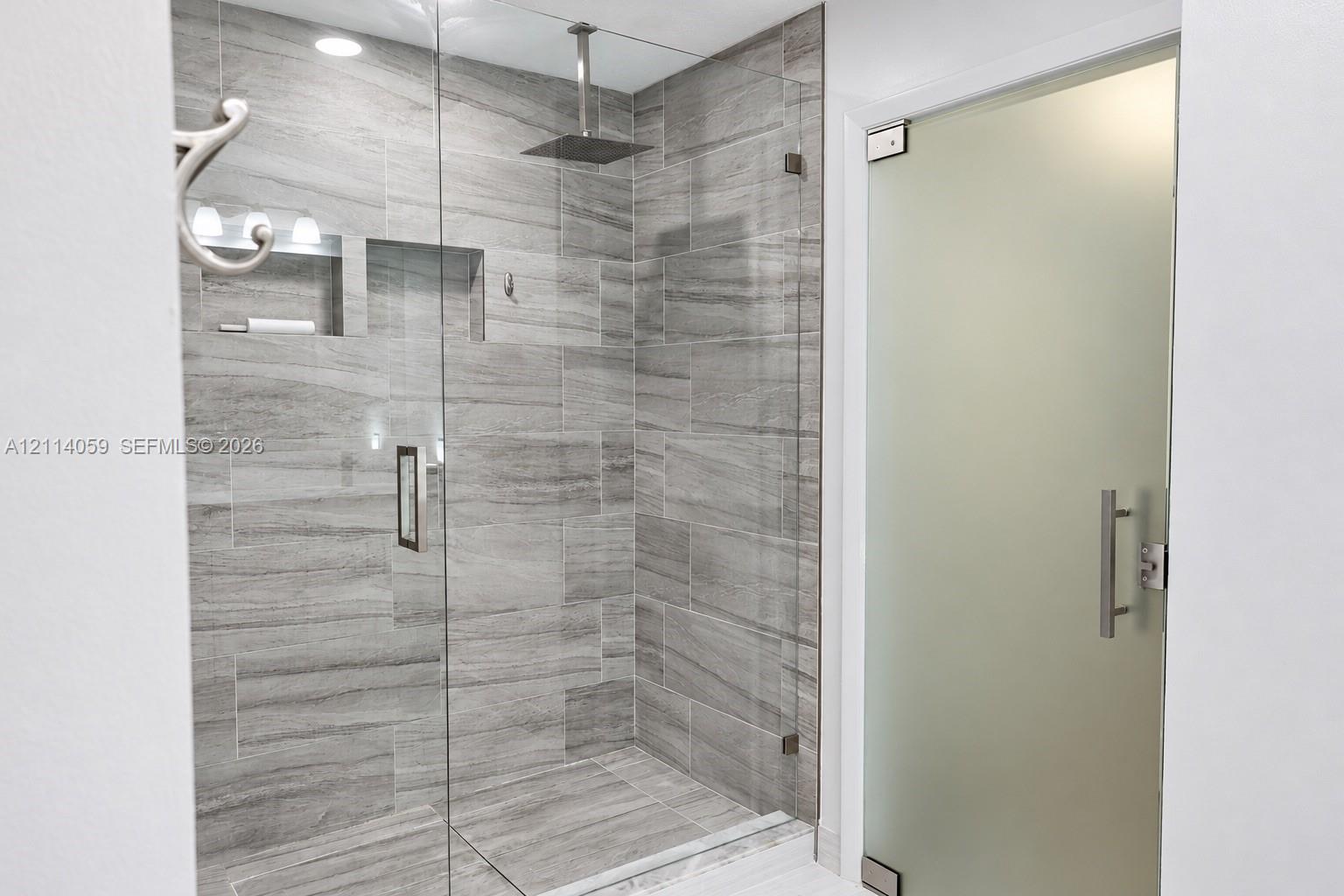 Walk-in shower featuring floor-to-ceiling stone-pattern tile, a ceiling-mounted rainfall showerhead, recessed shelving, and a frameless glass enclosure with a frosted glass entry door