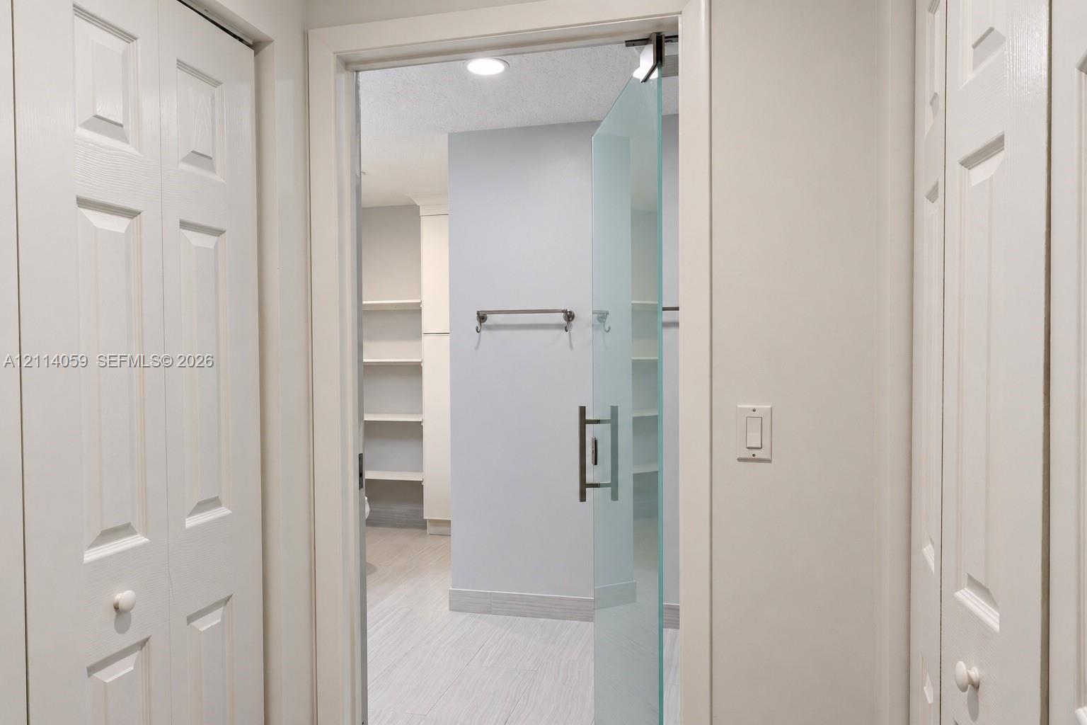 Frameless frosted glass door with a brushed metal handle, recessed ceiling lighting, built-in open shelving, and dual white paneled folding doors