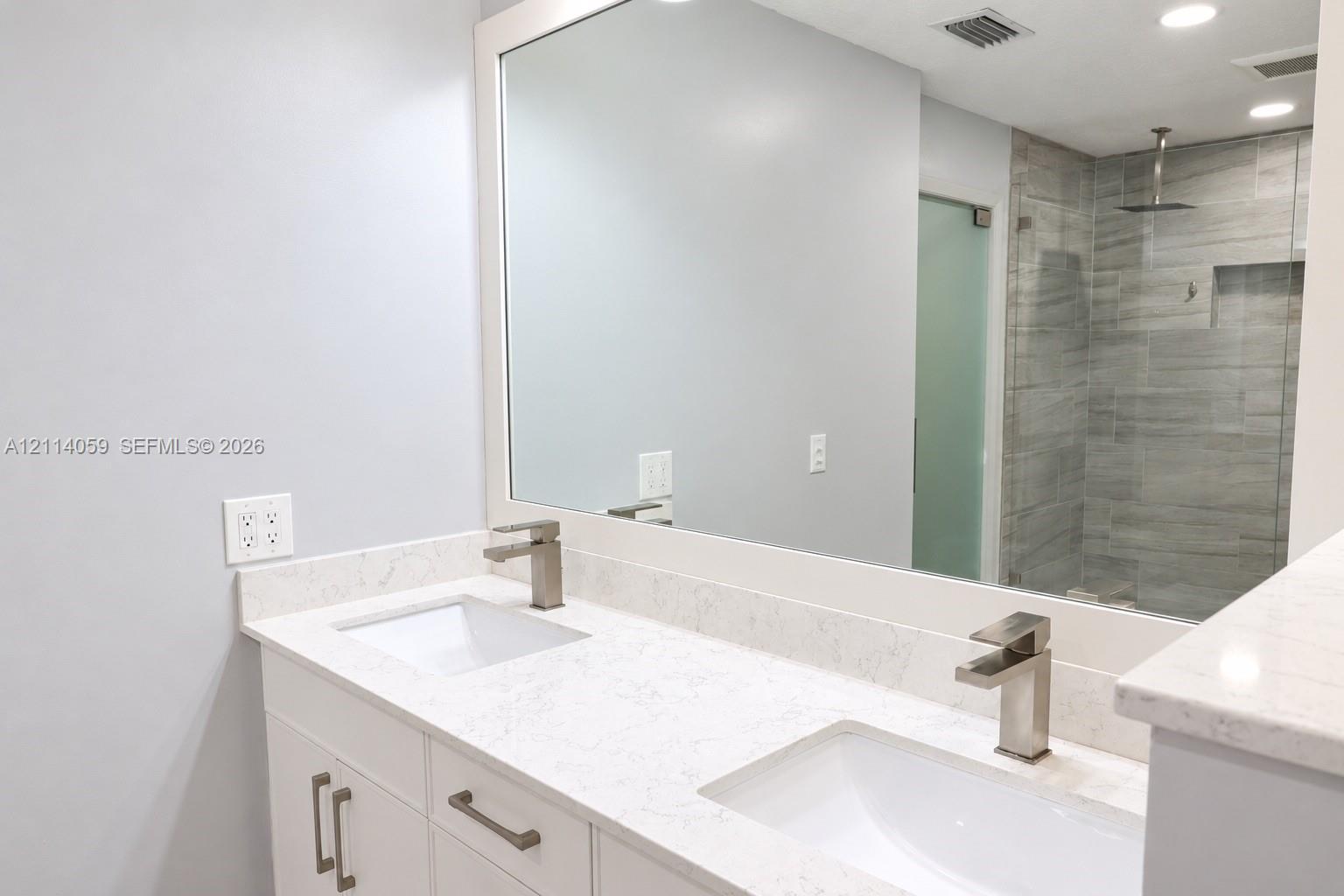 Double vanity featuring white cabinetry, stone-finish countertops, rectangular undermount sinks, and brushed-finish faucets, paired with a walk-in shower encased in glass with tile wall surfacing and a built-in recessed shelf
