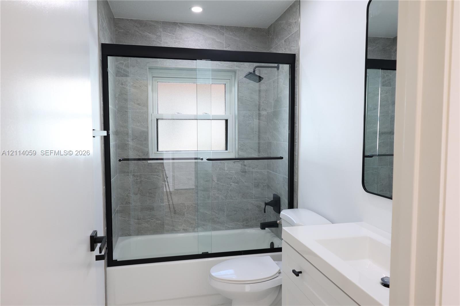 Bathtub enclosure with glass sliding doors, matte black fixtures, full-height stone-finish tile surround, recessed lighting, and a white vanity with an integrated sink