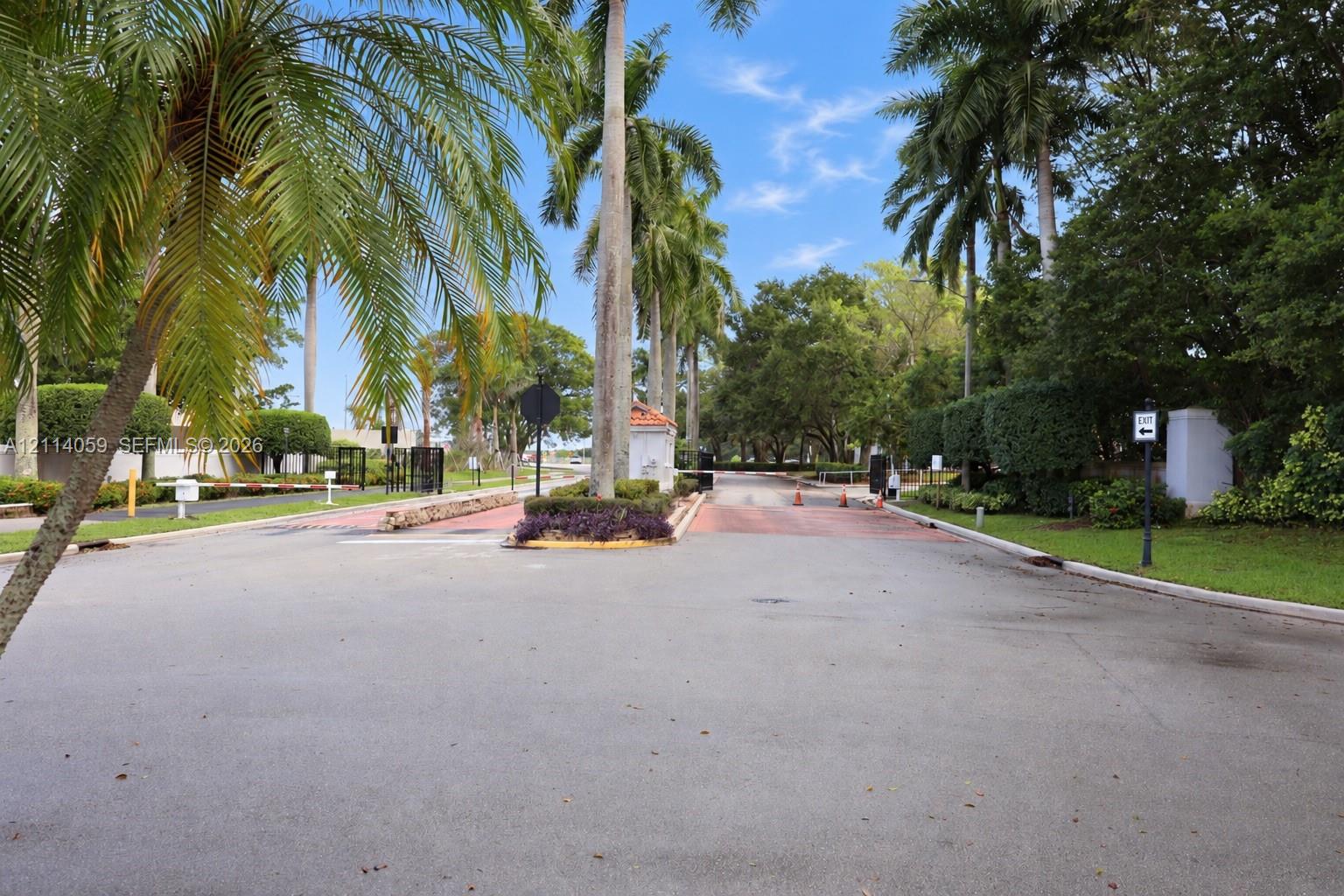 Gated entrance featuring a paved roadway, manicured landscaping, and mature palm trees