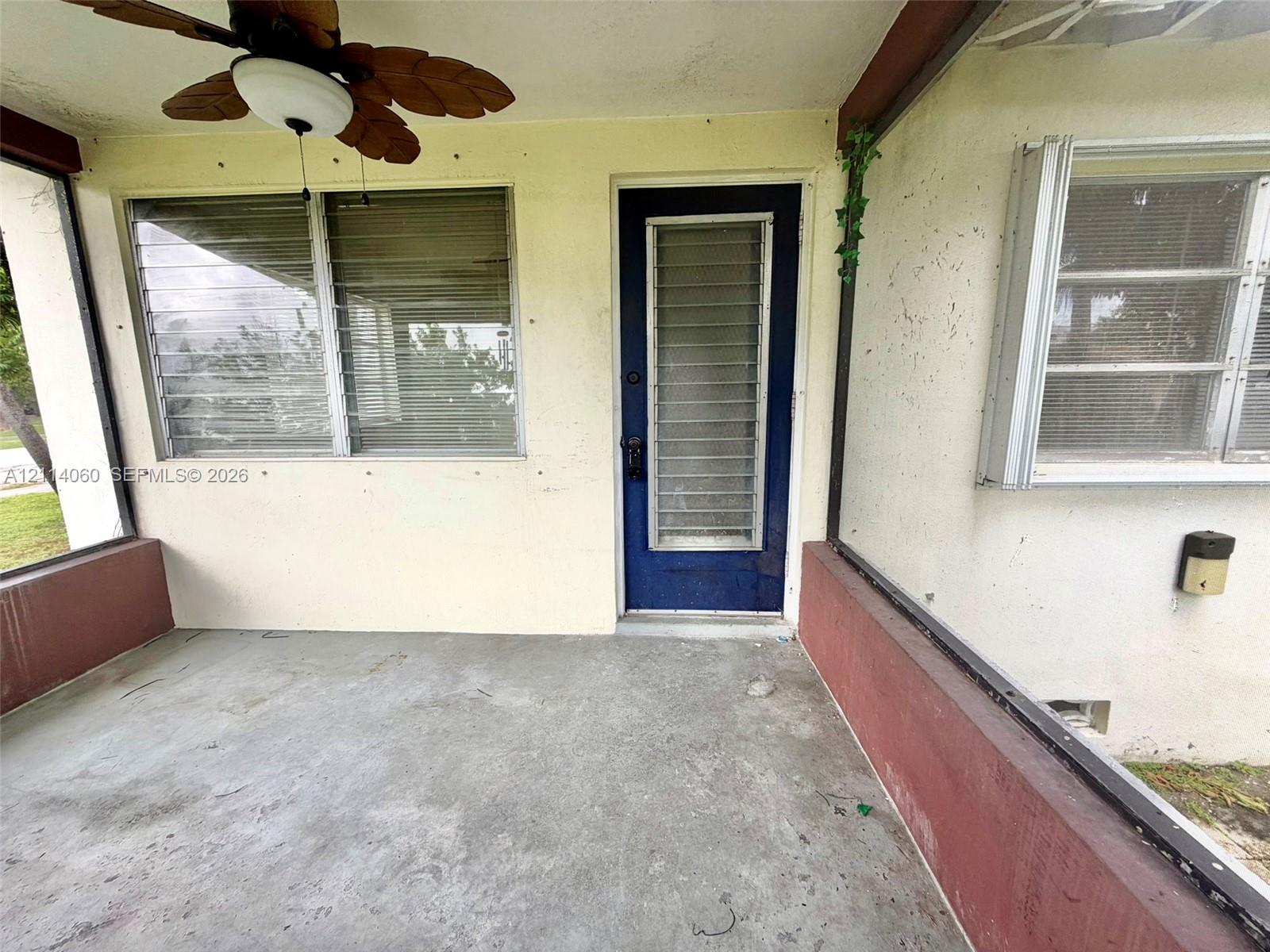 Screened-in porch featuring a concrete floor, ceiling fan with palm-leaf style blades, louvered windows, and a metal-frame entrance door with integrated ventilation slats
