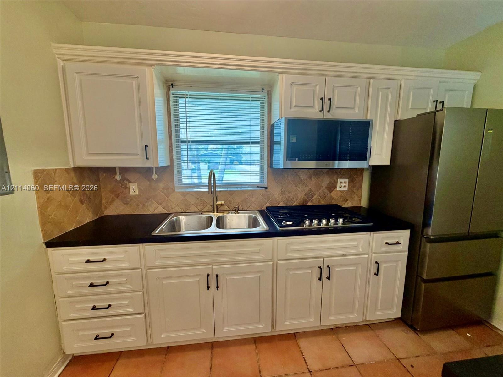 Kitchen featuring white shaker-style cabinetry, dark stone countertops, a dual-basin stainless steel sink, a built-in stainless steel gas cooktop, and a stone tile backsplash