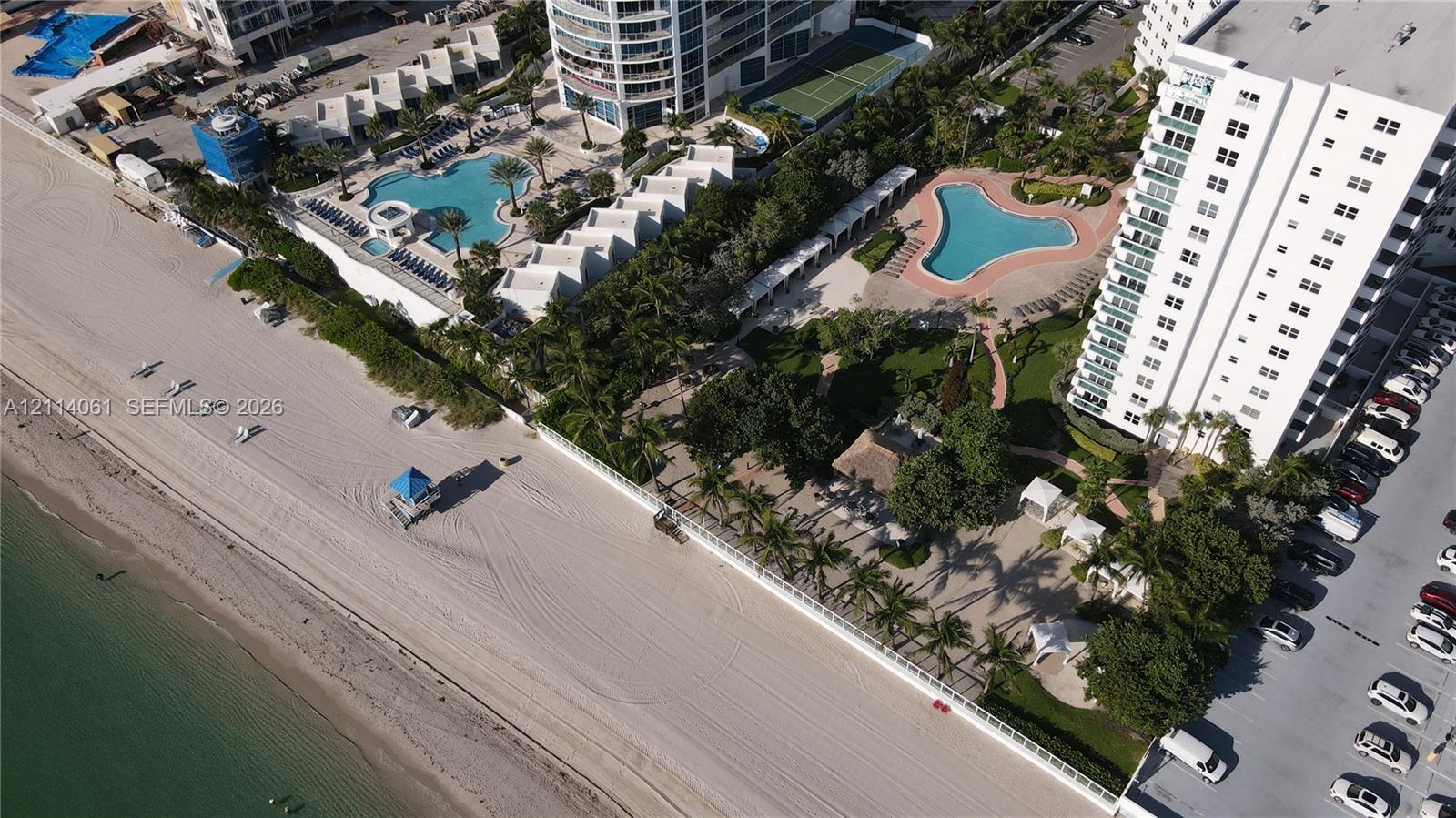 Aerial perspective of beachfront property featuring direct ocean access, multiple swimming pools, lush landscaped grounds with mature palm trees, and gated perimeters