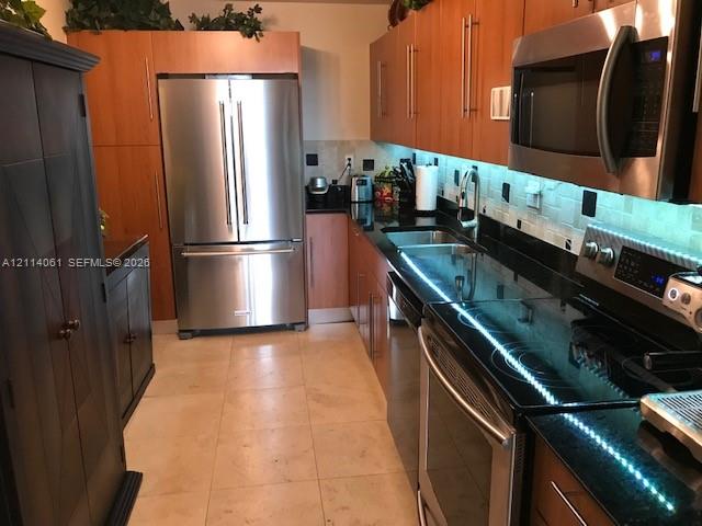 Kitchen featuring natural wood-finish cabinetry, dark solid-surface countertops, light stone tile flooring, under-cabinet lighting, and stainless steel appliances
