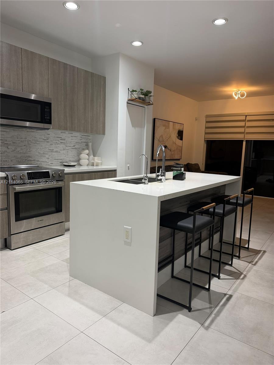 Kitchen island featuring a white countertop and stainless steel double-basin sink, wood-finish cabinetry, stainless steel appliances, stone-textured tile backsplash, and large-format tile flooring
