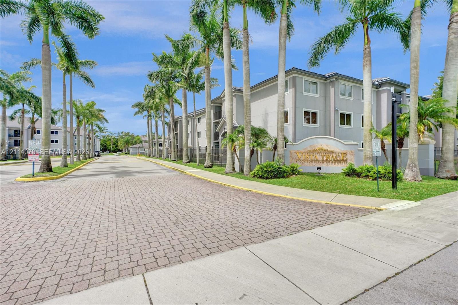 Multi-story residential buildings featuring light-toned masonry exteriors, paver-style roadway, mature palm trees, and manicured landscaping