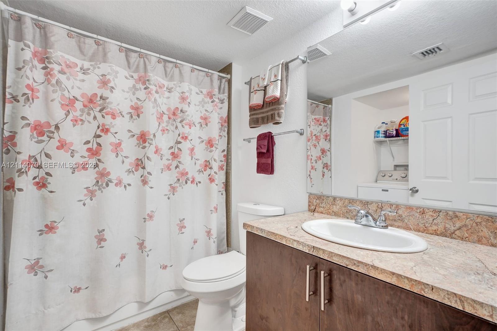 Bathroom vanity featuring a stone-patterned countertop, oval drop-in sink, and dark wood-finish cabinetry, complemented by a large wall-mounted mirror and a built-in laundry closet