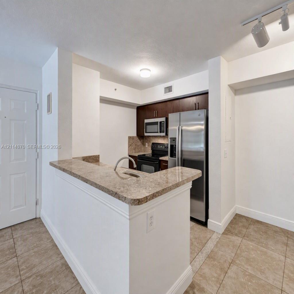 Kitchen featuring a peninsula with a stone-finish countertop, stainless steel refrigerator, built-in microwave, dark wood-finish cabinetry, and tile flooring