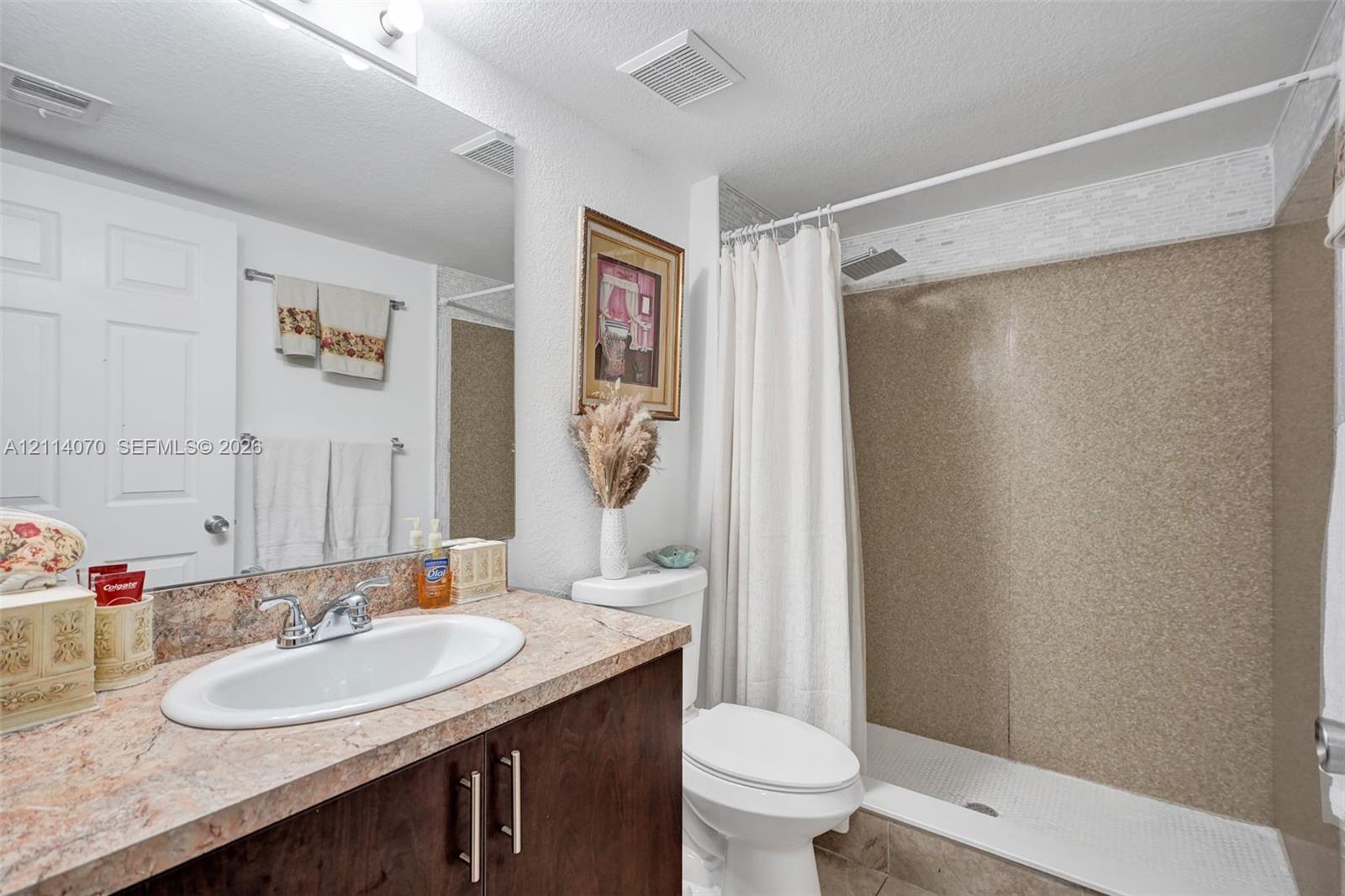 Bathroom featuring a vanity with a stone-finish countertop, oval drop-in sink, and dark wood-tone cabinetry, complemented by a walk-in shower with textured wall surround and an overhead showerhead