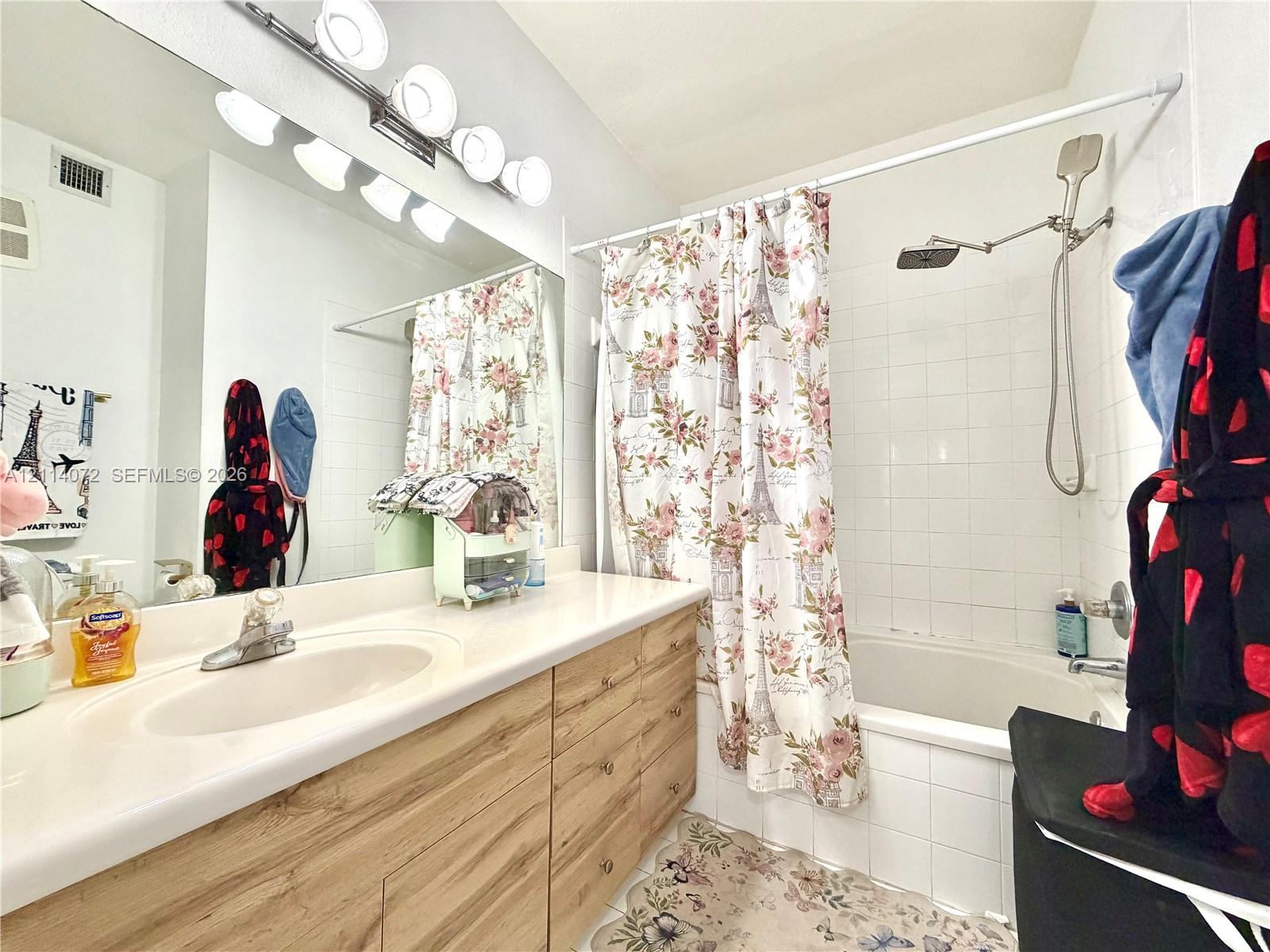 Bathroom featuring a vanity with light wood-finish cabinetry, an integrated sink with a polished chrome faucet, a wall-to-wall mirror, multi-bulb lighting, and a shower-tub combination with white wall tile