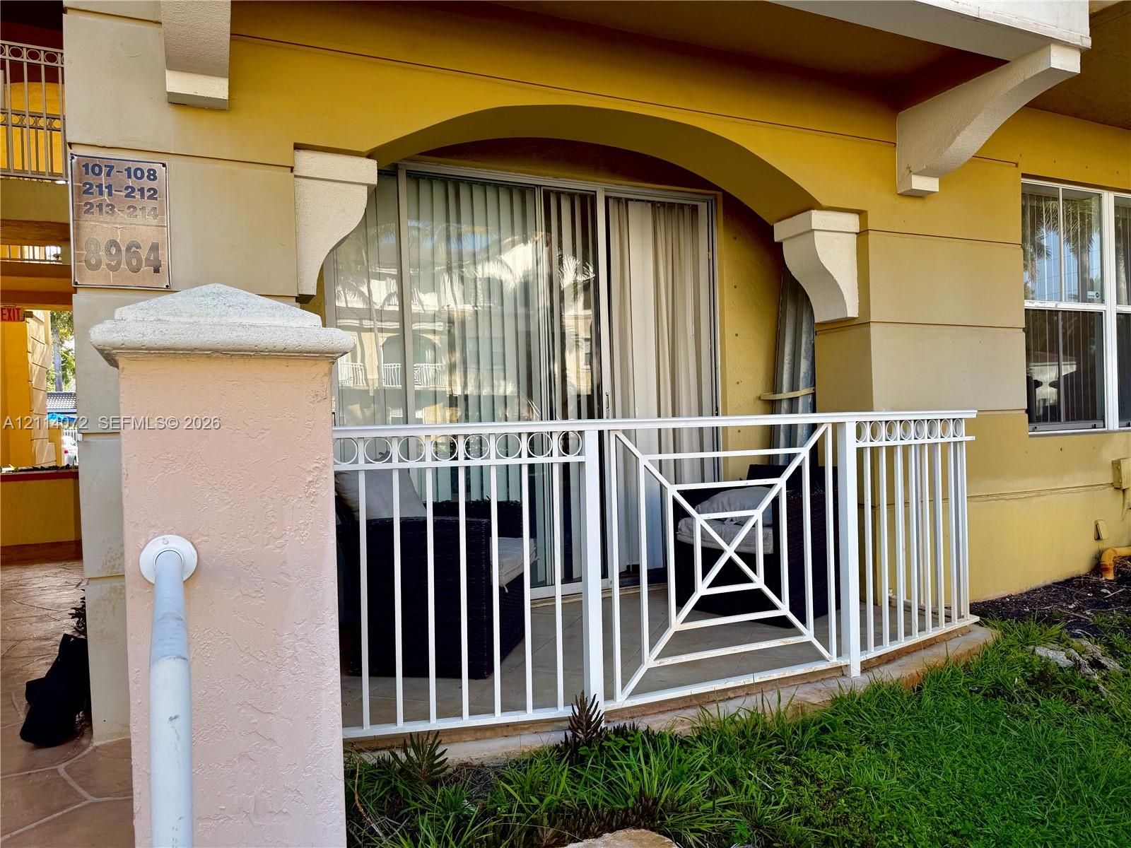 Covered ground-level patio featuring a white metal railing, arched stucco architectural detailing, and glass sliding doors