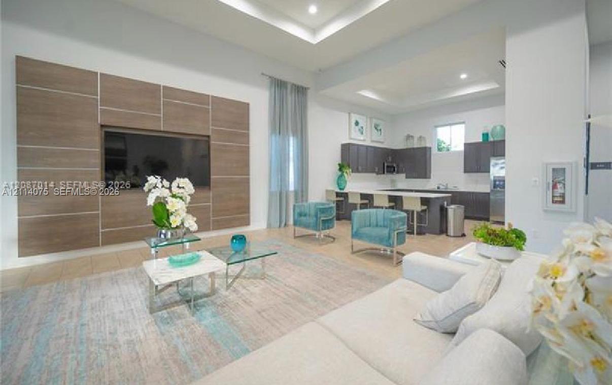 Living area featuring a custom wood-finish paneled accent wall, tray ceilings, recessed lighting, and kitchen with dark cabinetry and a central island