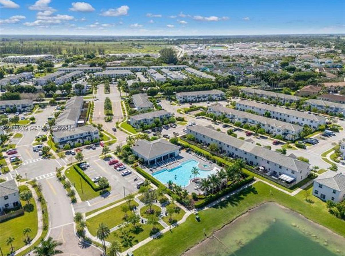 Aerial perspective of a multi-unit residential community featuring a centrally located swimming pool, paved surface parking areas, landscaped grounds, and a water feature