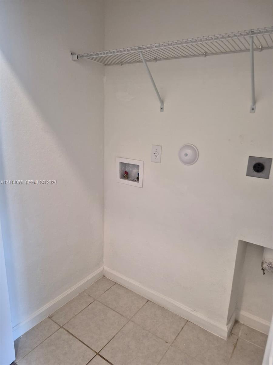 Utility space featuring ceramic tile flooring, a recessed water supply box, an electrical outlet, a dedicated power connection, and a mounted wire shelving unit