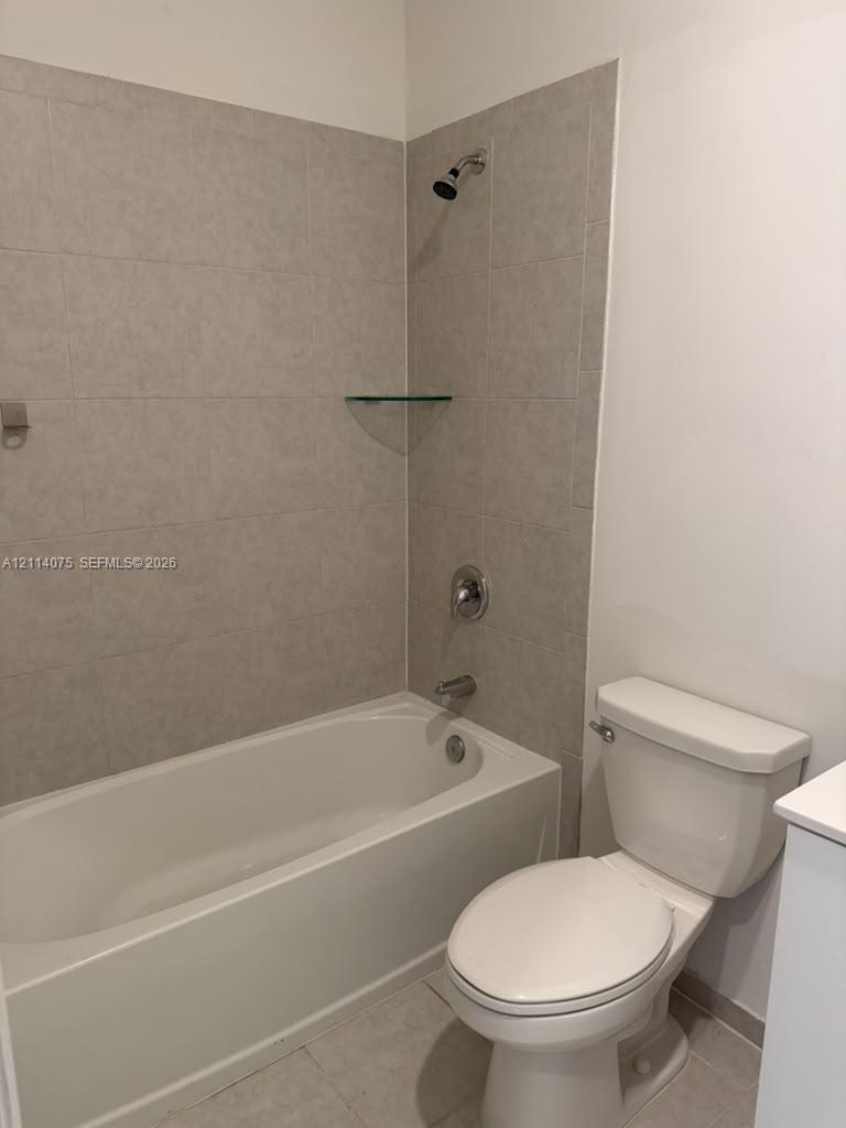 Full-size bathtub with integrated shower, neutral-toned wall tile surround, built-in glass corner shelving, chrome-finish plumbing fixtures, and standard-height porcelain commode