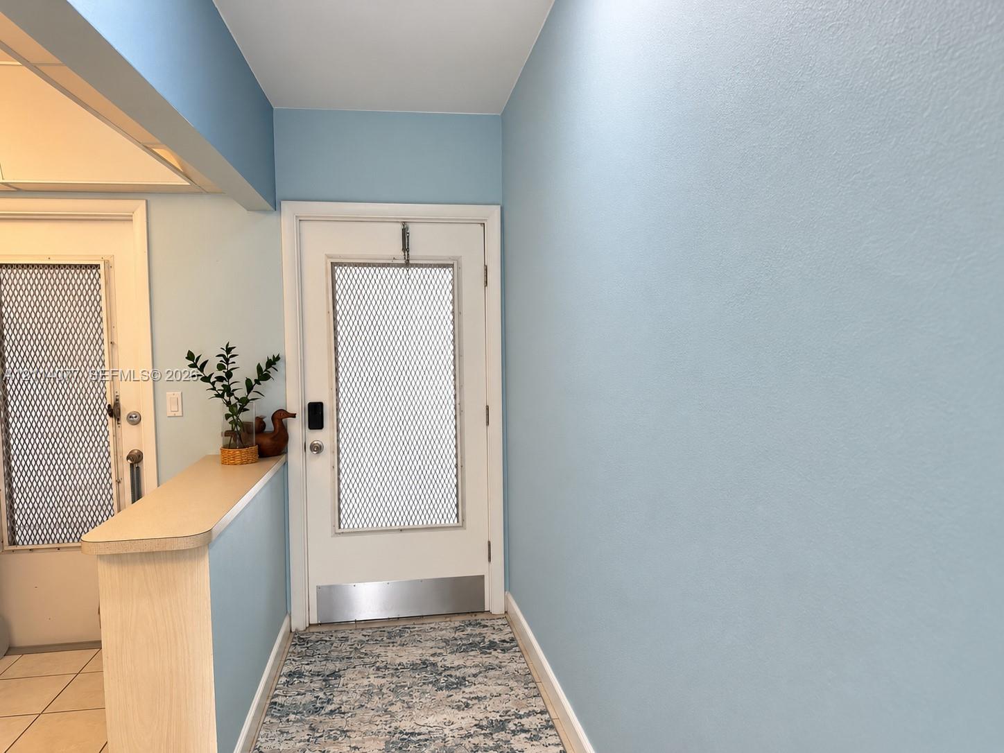 Entrance featuring a white door with a metal kickplate, decorative mesh insert, and integrated keypad lock, complemented by a solid-surface half-wall divider and overhead skylight