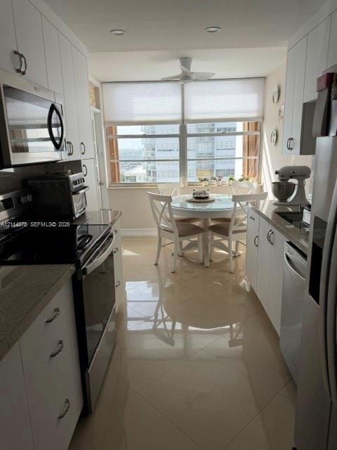 Galley kitchen featuring white shaker-style cabinetry, stone countertops, stainless steel appliances, large-format tile flooring, and oversized windows with window treatments