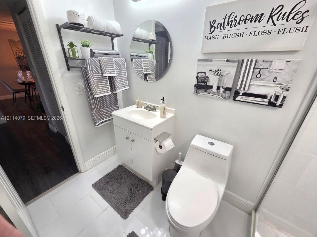 White vanity with integrated sink, circular wall-mounted mirror, white tile flooring, and two-piece elongated toilet