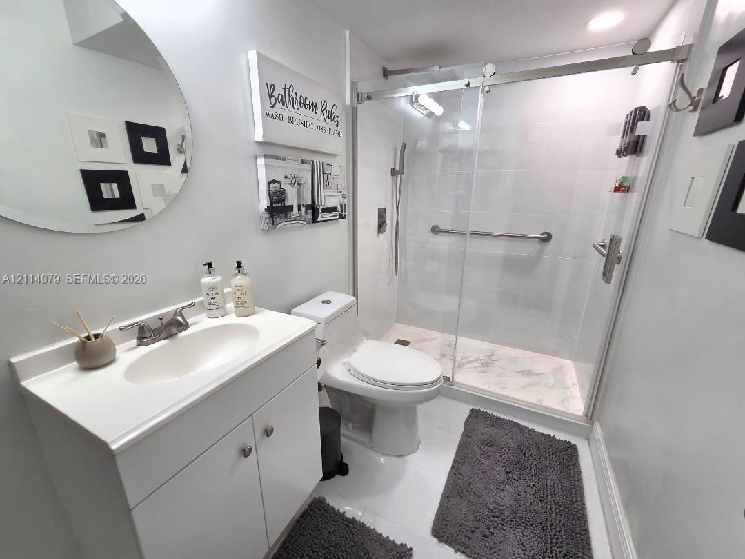 White vanity with integrated basin, walk-in shower featuring a glass sliding door, stone-patterned shower floor tile, and a circular wall-mounted mirror