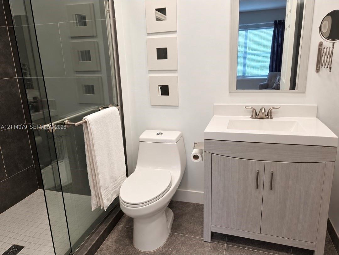 Bathroom featuring a wood-finish vanity with a rectangular sink, glass-enclosed shower with dark tile surround, and neutral floor tile