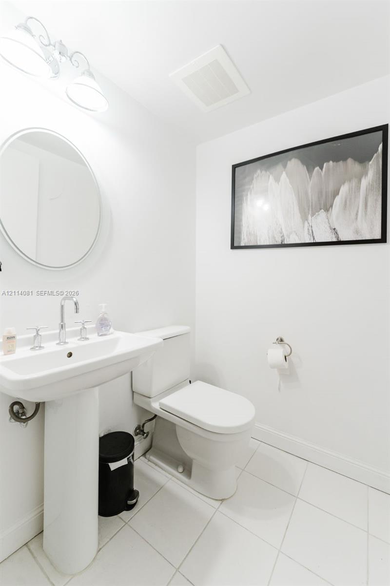 Pedestal sink featuring a polished chrome faucet, round wall-mounted mirror, ceramic tile flooring, and wall-mounted lighting fixture