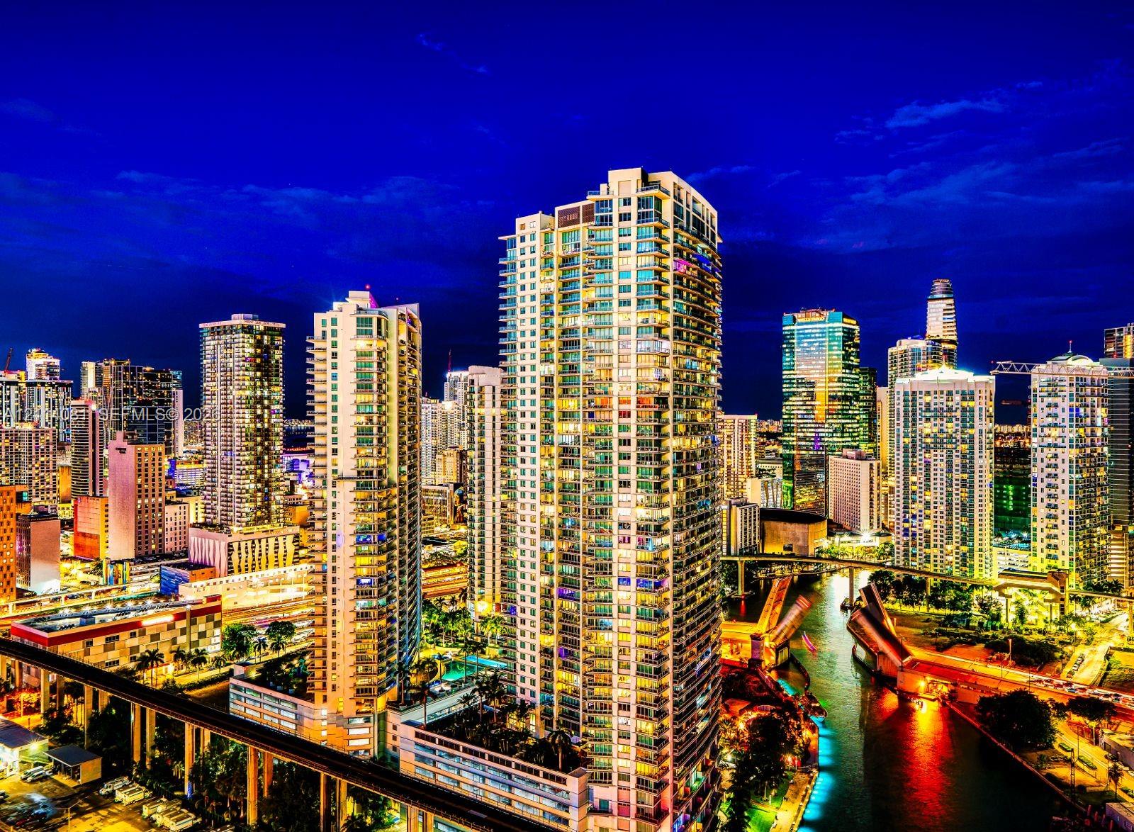 High-rise residential tower featuring glass-paneled balconies, floor-to-ceiling windows, and integrated exterior lighting, situated in a dense metropolitan center with a waterway