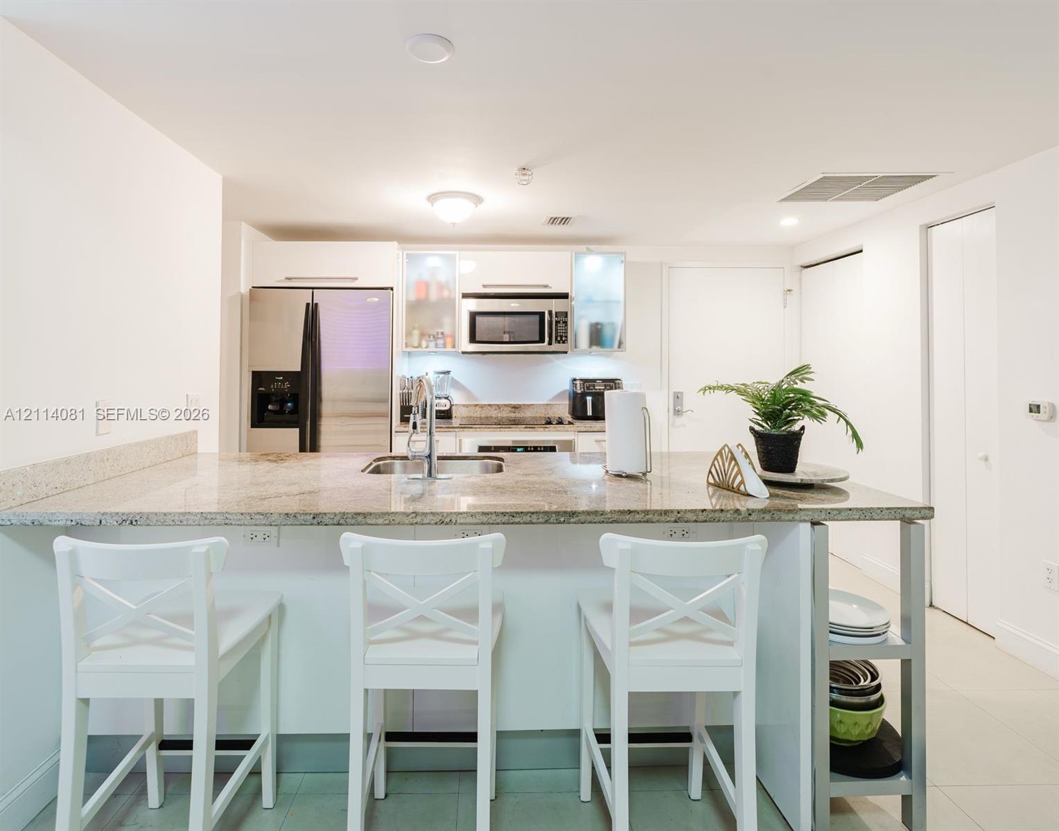 Kitchen featuring a stone-topped island with an integrated stainless steel sink, flat-panel white cabinetry, built-in stainless steel microwave, and stainless steel refrigerator