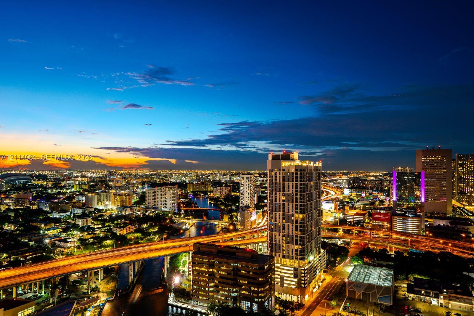 Aerial perspective featuring a high-rise structure, urban skyline, waterway, and elevated roadway infrastructure