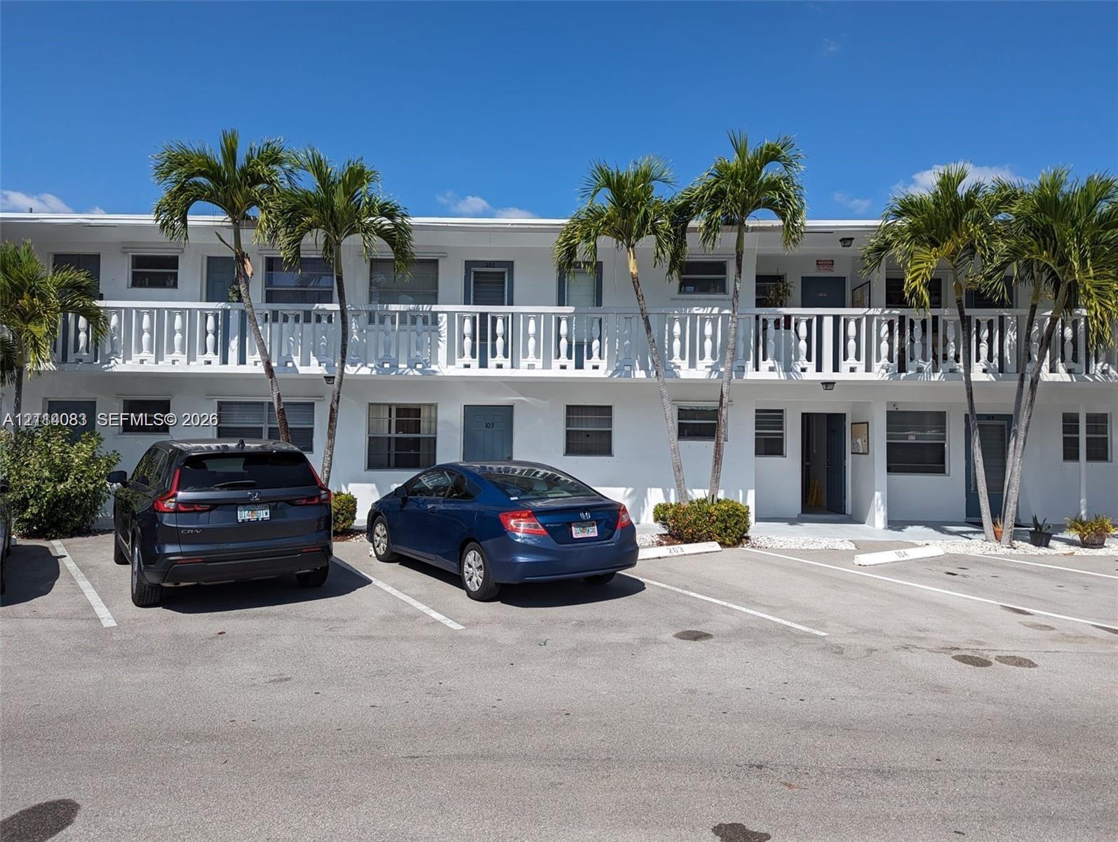 Two-story exterior featuring white stucco finish, second-level balcony with white balustrades, and asphalt surface parking