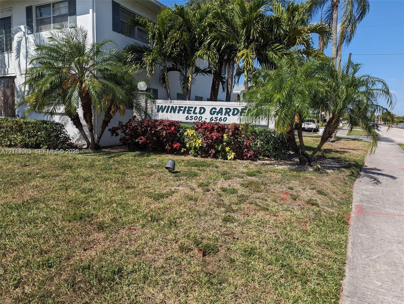 Exterior signage identifying Winfield Gardens, accompanied by mature palm trees, decorative shrubbery, and a concrete walkway