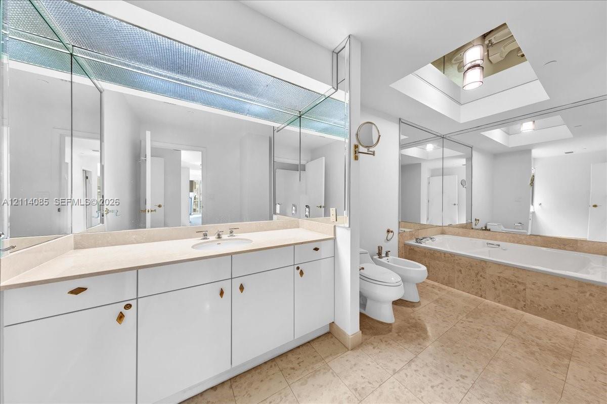 Bathroom featuring a white vanity with integrated stone countertop and sink, large wall-to-wall mirror, skylight, soaking tub with stone surround, and tile flooring