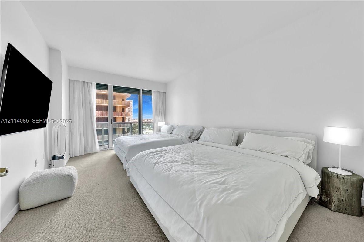 Floor-to-ceiling glass sliding doors opening to a private balcony, wall-mounted television, neutral carpeting, and smooth white wall finishes