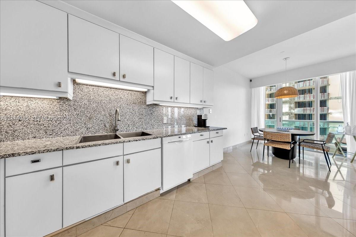 Kitchen featuring white cabinetry, stone-flecked countertops, full-height backsplash, polished tile flooring, and integrated dishwasher