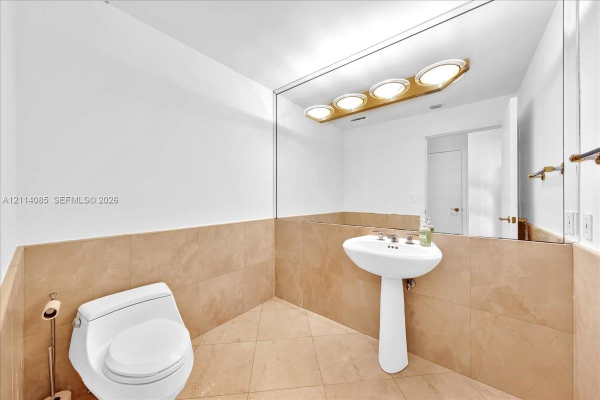 Pedestal sink with polished metal faucet, full-width wall mirror, four-bulb vanity light fixture, and large-format tile flooring and wainscoting