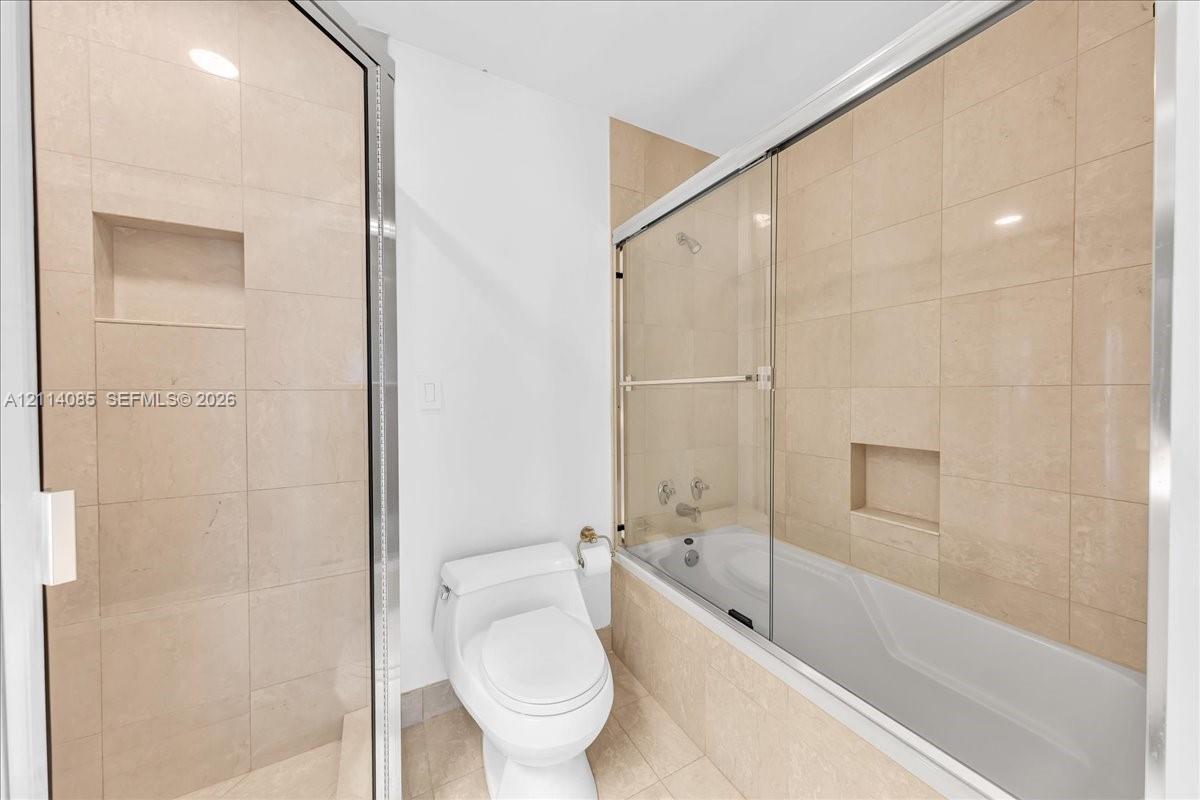 Neutral-toned tile surrounds in the shower and tub enclosure, glass sliding shower doors, built-in tile wall niches, and a white porcelain toilet