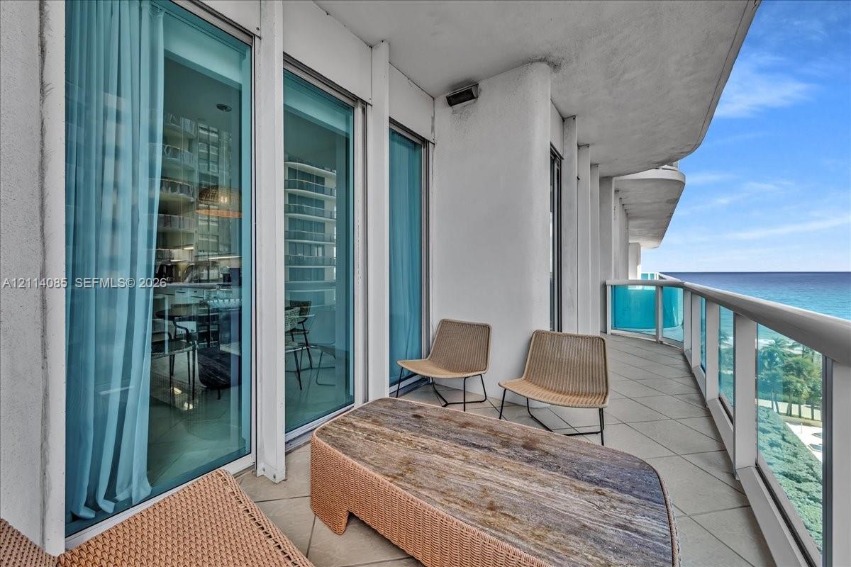 Spacious covered balcony with large-format tile flooring, floor-to-ceiling glass sliding doors, and a glass-paneled railing overlooking the ocean