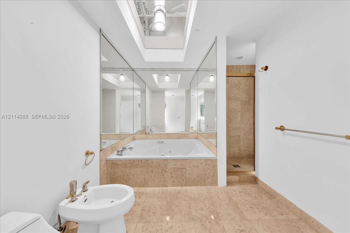 Jetted soaking tub with stone tile surround, skylight, wall-to-wall mirrors, pedestal sink, and separate walk-in shower with stone tile wall finishes