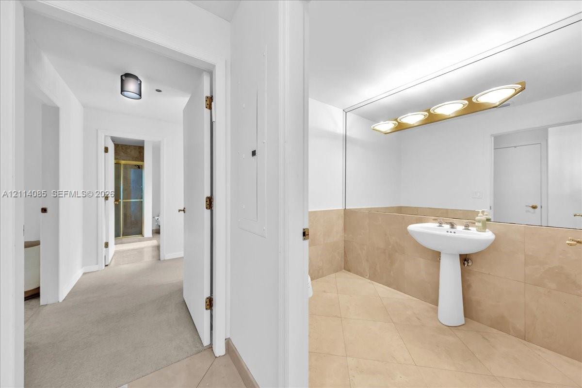 Bathroom featuring a pedestal sink, large vanity mirror with multi-bulb light fixture, beige tile flooring, and tile wall wainscoting