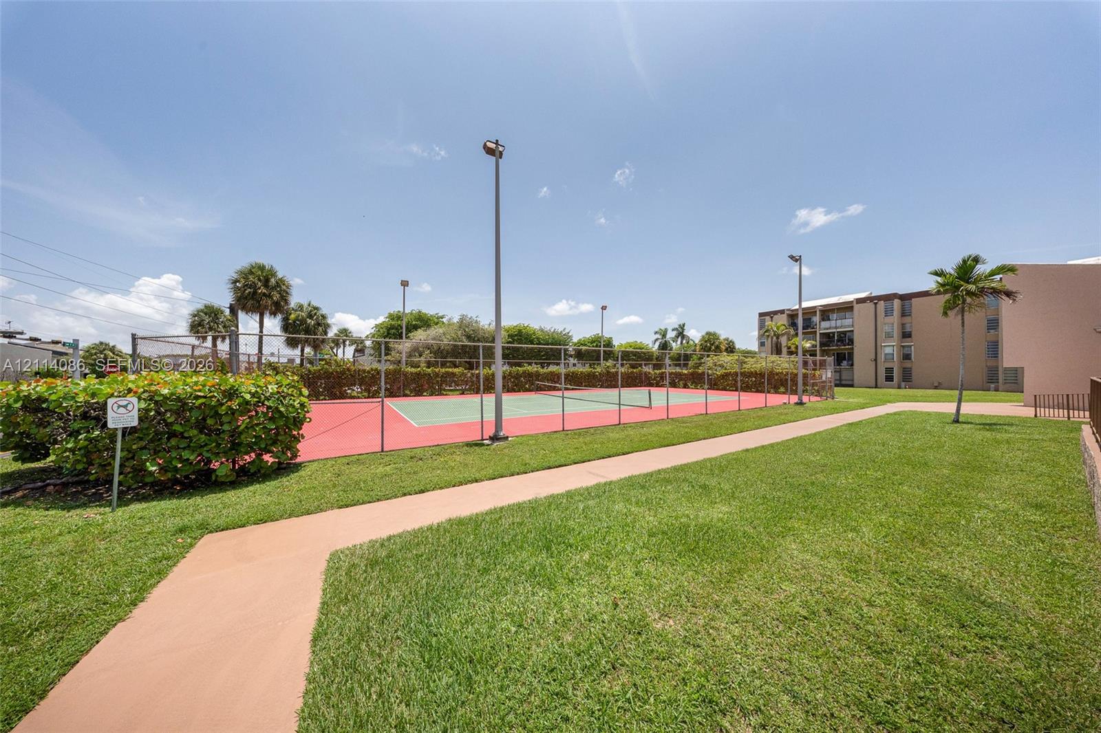Fenced tennis court with a red and green surface, surrounding perimeter lighting, and paved walkway set within a landscaped grass lawn