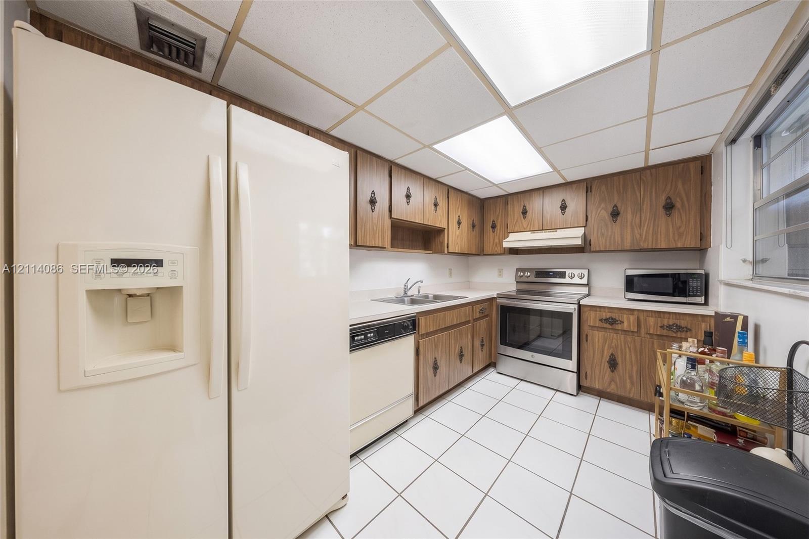 Kitchen featuring wood-finish cabinetry, stainless steel range, white tile flooring, drop ceiling panels, and ample solid-surface countertops