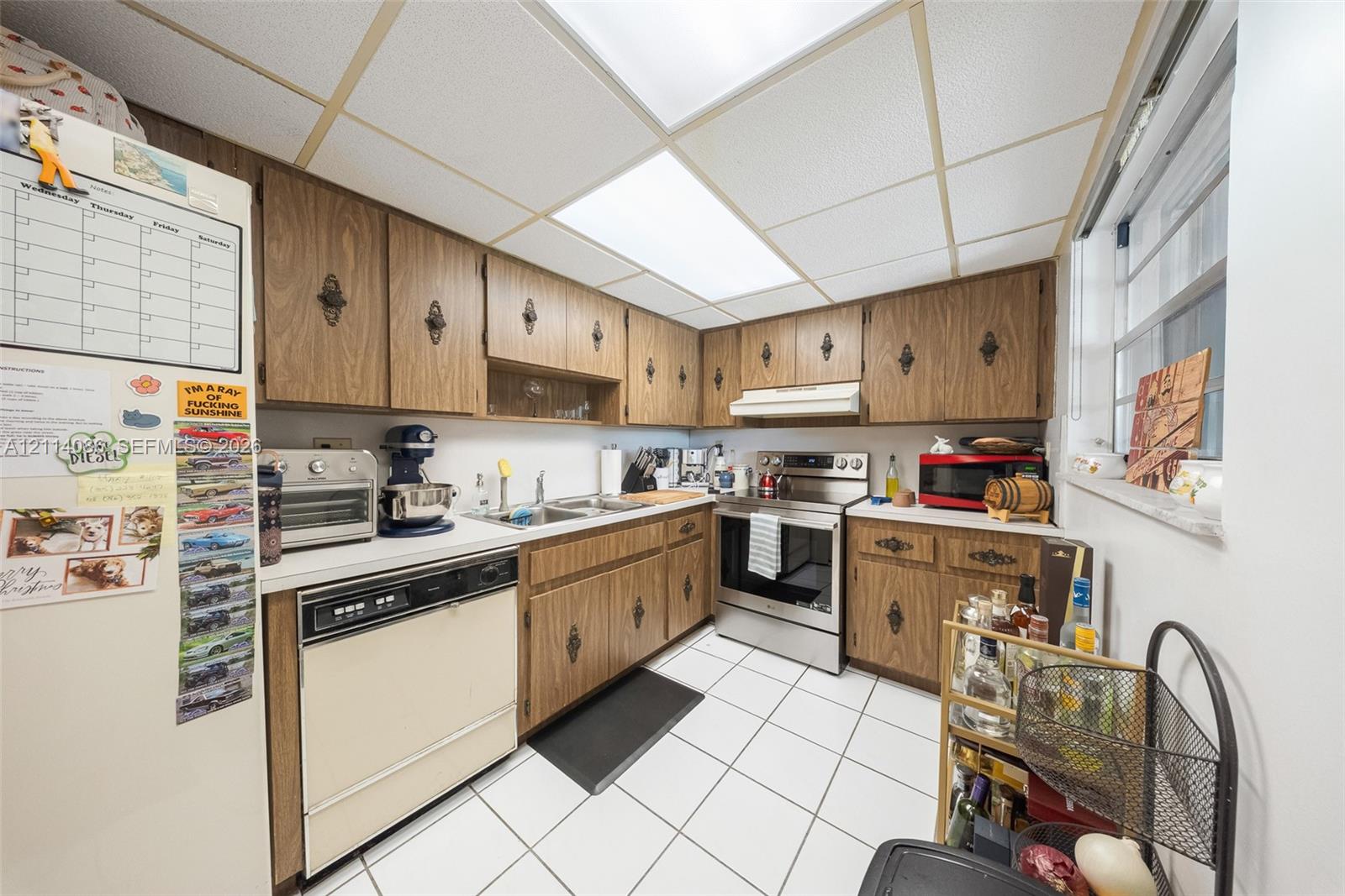 Kitchen featuring wood-finish cabinetry with ornate hardware, stainless steel slide-in range, integrated dishwasher, white tiled flooring, and drop-ceiling panel lighting