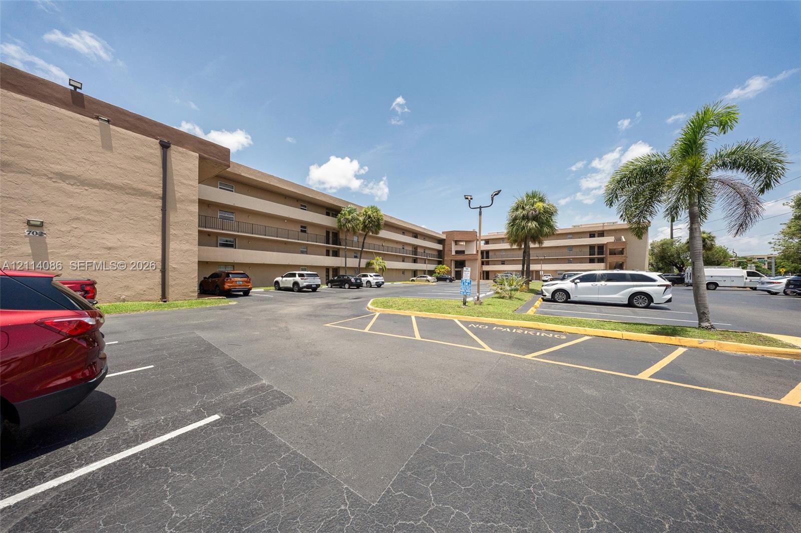 Multi-story exterior featuring a stucco facade, covered balconies, asphalt parking area, and mature palm trees