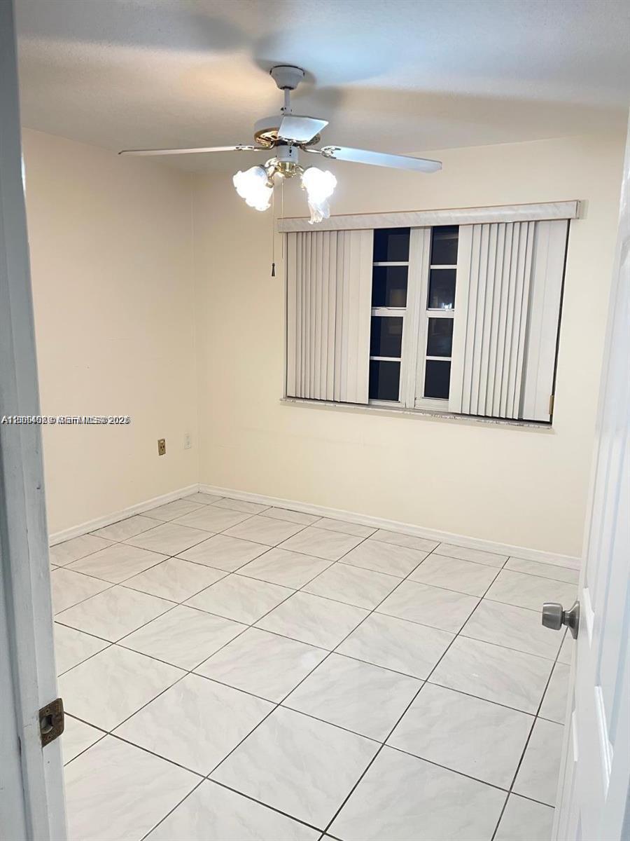 Neutral-toned interior featuring large-format tile flooring, a multi-blade ceiling fan with integrated lighting, and vertical window blinds Neutral-toned interior featuring large-format tile flooring, a multi-blade ceiling fan with integrated lighting, and vertical window blinds