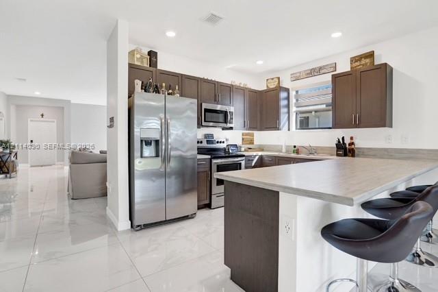 Kitchen featuring dark-finish cabinetry, stainless steel appliances, a peninsula with a stone-finish countertop, and light-toned tile flooring Kitchen featuring dark-finish cabinetry, stainless steel appliances, a peninsula with a stone-finish countertop, and light-toned tile flooring