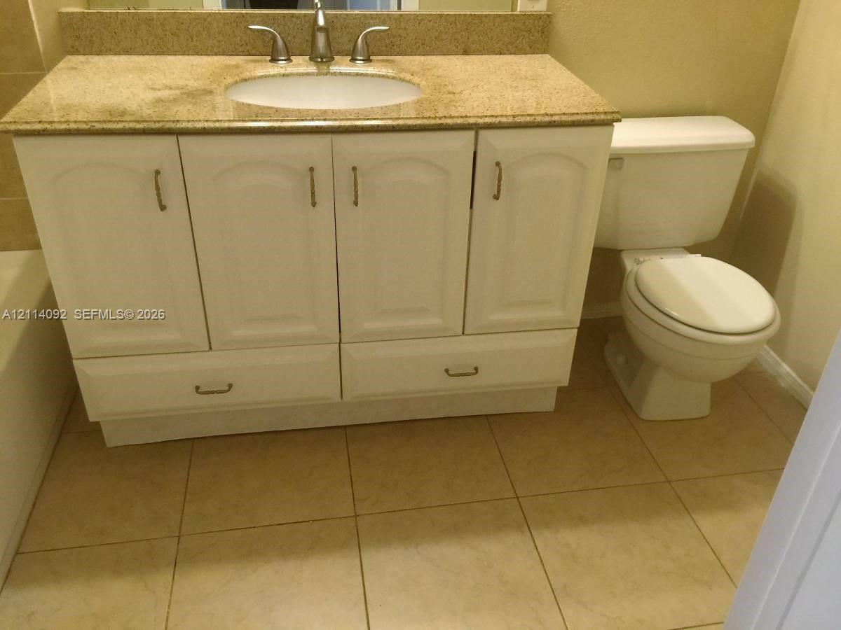 Vanity cabinet with white-finish cabinetry and stone-composite countertop, undermount sink with metal faucet, and ceramic tile flooring