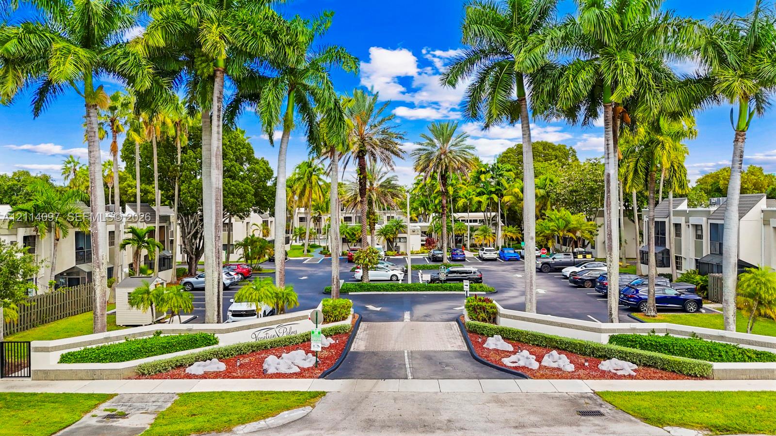 Gated community entrance featuring masonry pillars, manicured boxwood hedges, mature palm trees, and asphalt driveway Gated community entrance featuring masonry pillars, manicured boxwood hedges, mature palm trees, and asphalt driveway