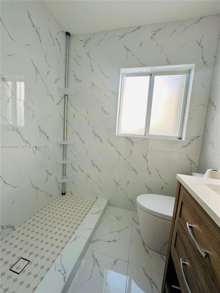 Walk-in shower featuring floor-to-ceiling large-format tile with grey veining, a geometric mosaic tile shower floor, a light-toned wood-finish vanity with a solid-surface countertop, and a sliding window